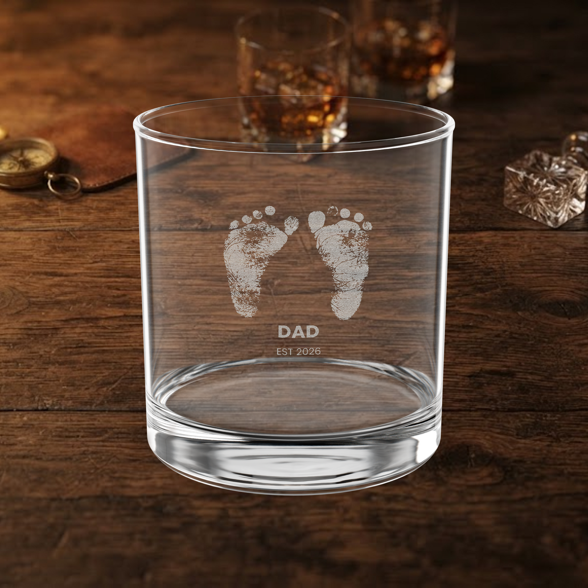 A clear glass rocks tumbler with etched baby footprints and the text 'DAD EST 2026' on a dark wooden table, with blurred whiskey glasses in the background.