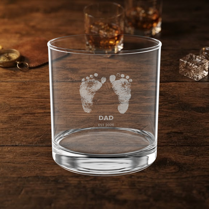 A clear glass rocks tumbler with etched baby footprints and the text 'DAD EST 2026' on a dark wooden table, with blurred whiskey glasses in the background.